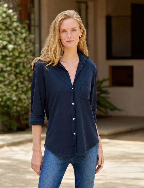 A woman wearing a dark navy blue button-down shirt with rolled sleeves and blue jeans, standing outdoors in front of a neutral-toned building., front view