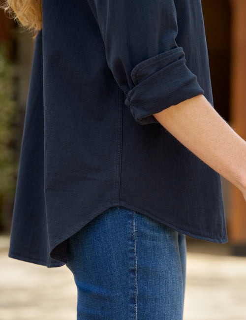 Close-up of a person wearing a dark navy blue long-sleeve top with rolled sleeves and blue jeans, cropped from the side against a softly blurred outdoor background., view 3