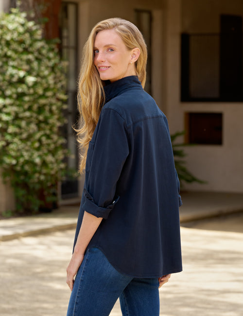 A woman with blonde hair wearing a dark navy blue button-up shirt and blue jeans, standing in an outdoor patio setting., alternate view