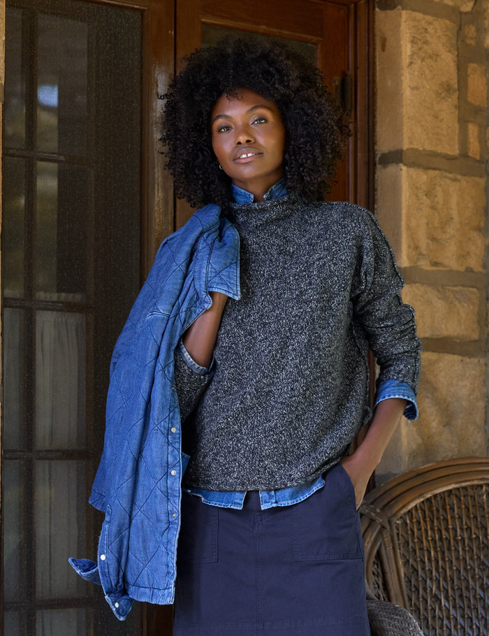 A woman stands on a stone porch wearing a dark gray textured mockneck sweater, a navy blue skirt, a blue denim jacket draped over her shoulder, and brown loafers. A wicker chair with a blue throw blanket is beside her.