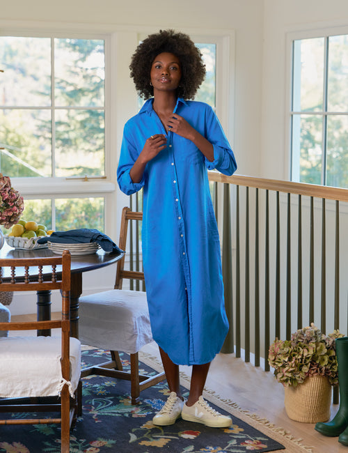 Woman wearing a blue dress standing in a room with large windows and furniture., front view