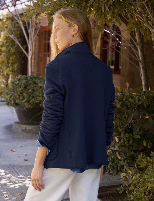 A woman wearing a navy blue jacket and white pants, photographed from behind in an outdoor setting with trees and a building in the background., alternate view