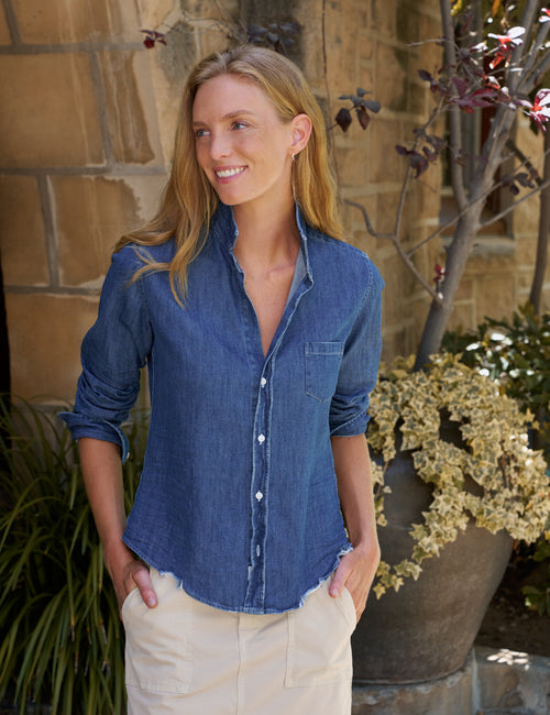 A woman wearing a blue denim button-down shirt and beige skirt, standing outdoors in front of a stone wall with potted plants., front view