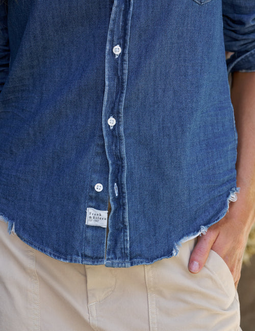 Close-up of a person wearing a blue denim button-up shirt with a raw, frayed hem and light beige skirt., view 3