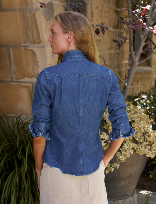 A woman viewed from behind wearing a blue denim button-up shirt with rolled sleeves and light beige skirt, standing outdoors in front of a stone wall and potted plants., alternate view