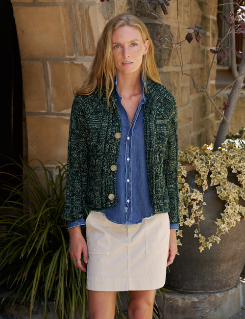 A woman stands outdoors in front of a stone wall wearing a dark green textured knit cardigan over a blue button-up shirt and a light beige mini skirt., front view