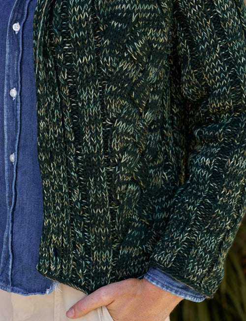 Close-up of a person wearing a dark green cable-knit sweater over a blue button-up shirt and beige skirt, with one hand in a pocket., view 3