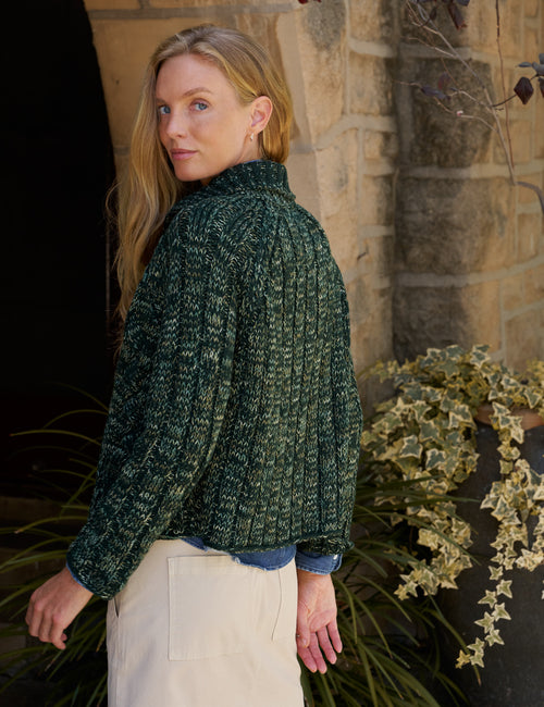 A woman wearing a dark green textured cable-knit mock-neck sweater and cream skirt, standing outdoors in front of a stone wall and green plants., alternate view