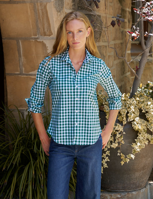 A woman wearing a green and white gingham button-down shirt with rolled-up sleeves and blue jeans, standing outdoors in front of a stone wall and potted plants., front view