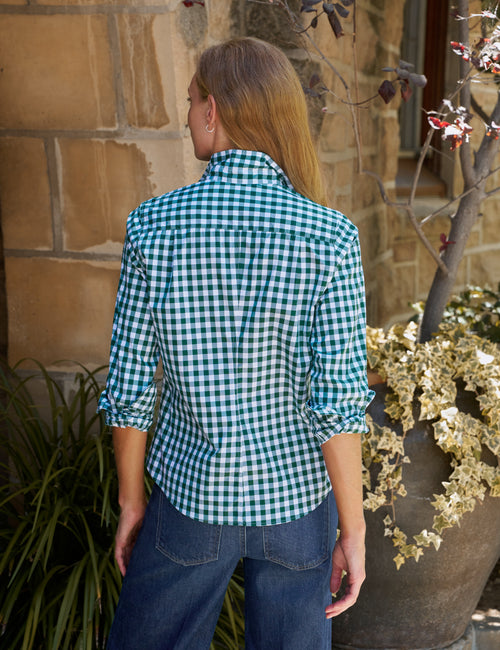 A woman viewed from behind wearing a green and white gingham long-sleeve button-down shirt and dark blue jeans, standing outdoors in front of a stone wall., alternate view