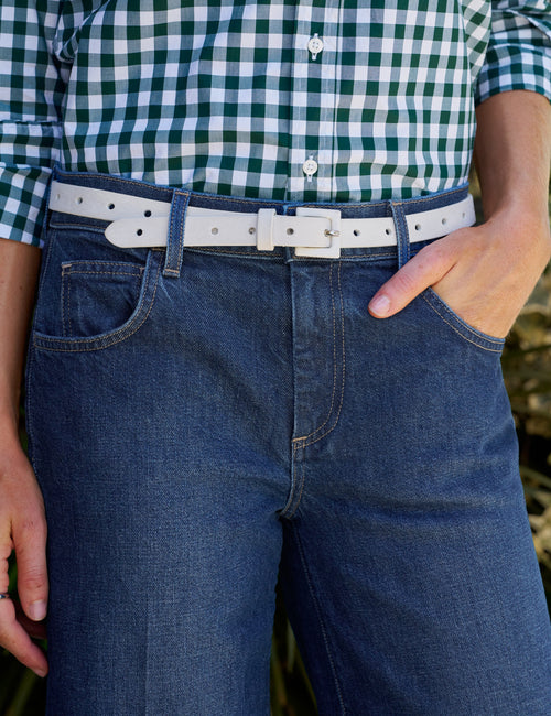 A close-up of a person wearing a white leather belt, styled with dark blue jeans and a green and white gingham shirt., front view