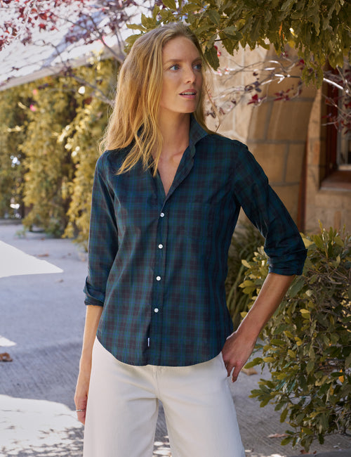 A woman wearing a dark teal and green plaid button-down shirt with white pants, standing outdoors in front of a stone wall and green foliage., front view