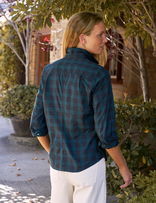 A woman wearing a dark teal and green plaid button-down shirt with white pants, photographed outdoors in a garden setting., alternate view