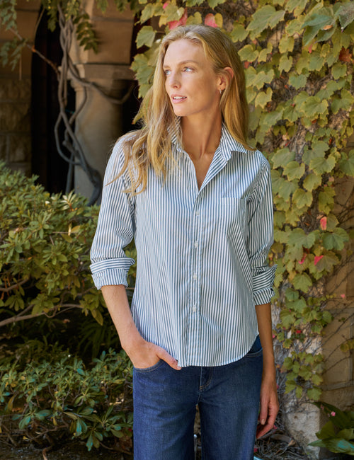 A woman wearing a green and white vertically striped button-down shirt with dark blue jeans, standing outdoors in front of a stone wall covered in green ivy., front view
