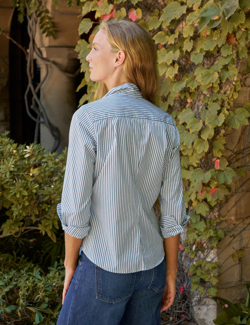A woman shown from the back wearing a green and white vertically striped button-down shirt with rolled-up sleeves, paired with dark blue jeans, standing outdoors against a background of green ivy and a stone wall., alternate view