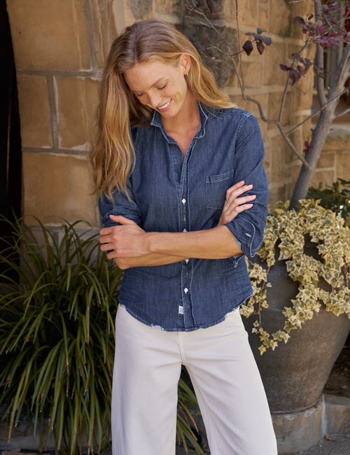A woman wearing a dark blue denim button-up shirt and off-white pants, standing outdoors in front of a stone wall with plants., front view