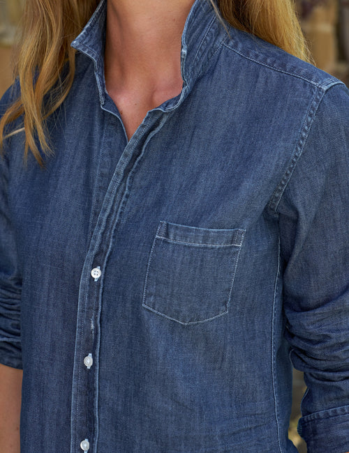 Close-up of a person wearing a blue denim button-up shirt with a chest pocket, shown from the neck to the waist against a softly blurred outdoor background., view 3