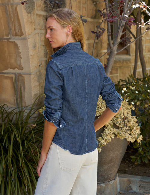 A woman wearing a blue denim button-up shirt and off-white pants, photographed from the back in an outdoor setting with a stone wall and potted plants., alternate view