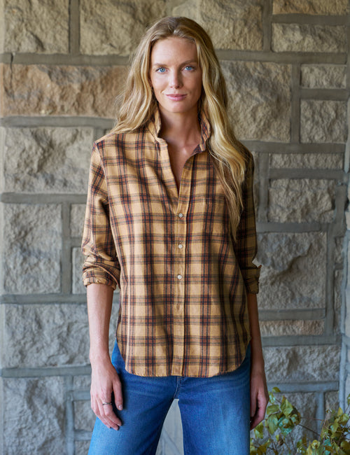 A woman with long blonde hair stands in front of a stone wall wearing a tan and brown plaid button-up shirt and blue jeans., front view