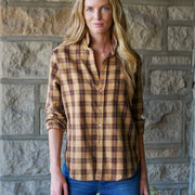 A woman with long blonde hair stands in front of a stone wall wearing a tan and brown plaid button-up shirt and blue jeans.