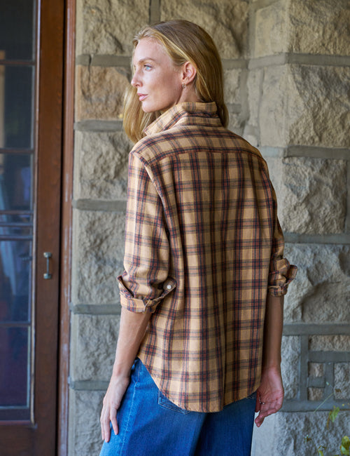A woman wearing a tan and brown plaid flannel shirt with rolled-up sleeves and blue jeans, standing in front of a stone wall., alternate view
