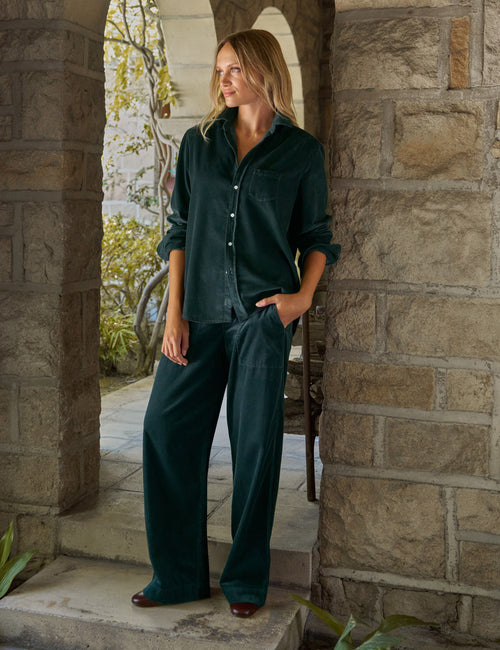 A woman stands in a stone archway doorway wearing a matching dark green corduroy button-up shirt and wide-leg pants set., front view