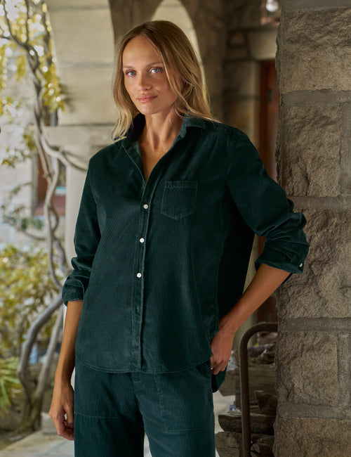 A woman wearing a dark teal-green corduroy button-up shirt and matching corduroy pants, standing in a stone-walled outdoor corridor., front view