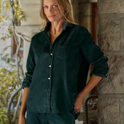 A woman wearing a dark teal-green corduroy button-up shirt and matching corduroy pants, standing in a stone-walled outdoor corridor.