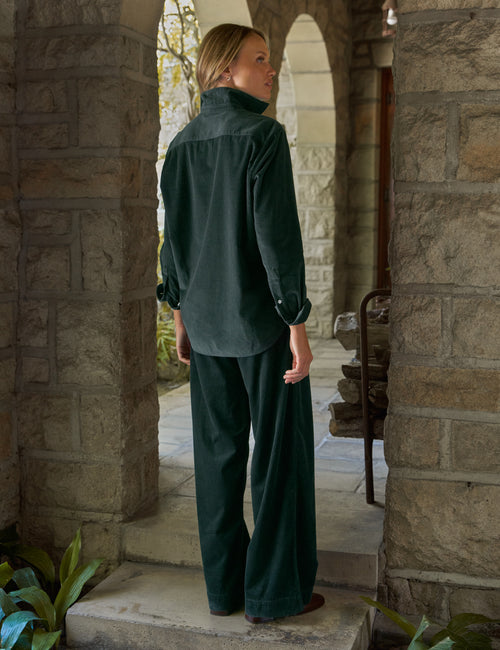 A woman wearing a dark forest green corduroy long-sleeve shirt and matching wide-leg trousers, standing in a stone archway doorway., alternate view