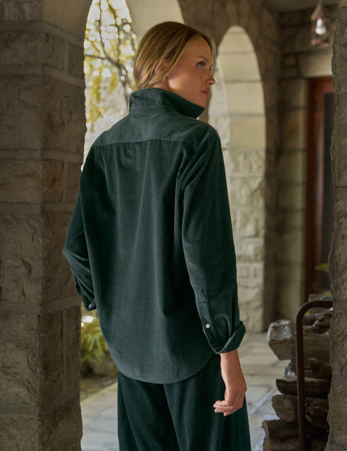 A woman wearing a dark forest green corduroy shirt and matching pants, standing in a stone archway corridor., alternate view