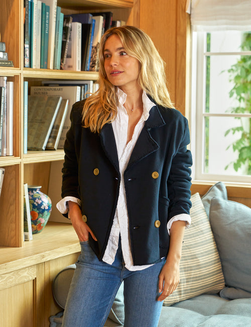 A woman standing in a home setting, wearing a navy blue mini Belfast sweatshirt peacoat with luxurious horn buttons over a white shirt and blue jeans., front view