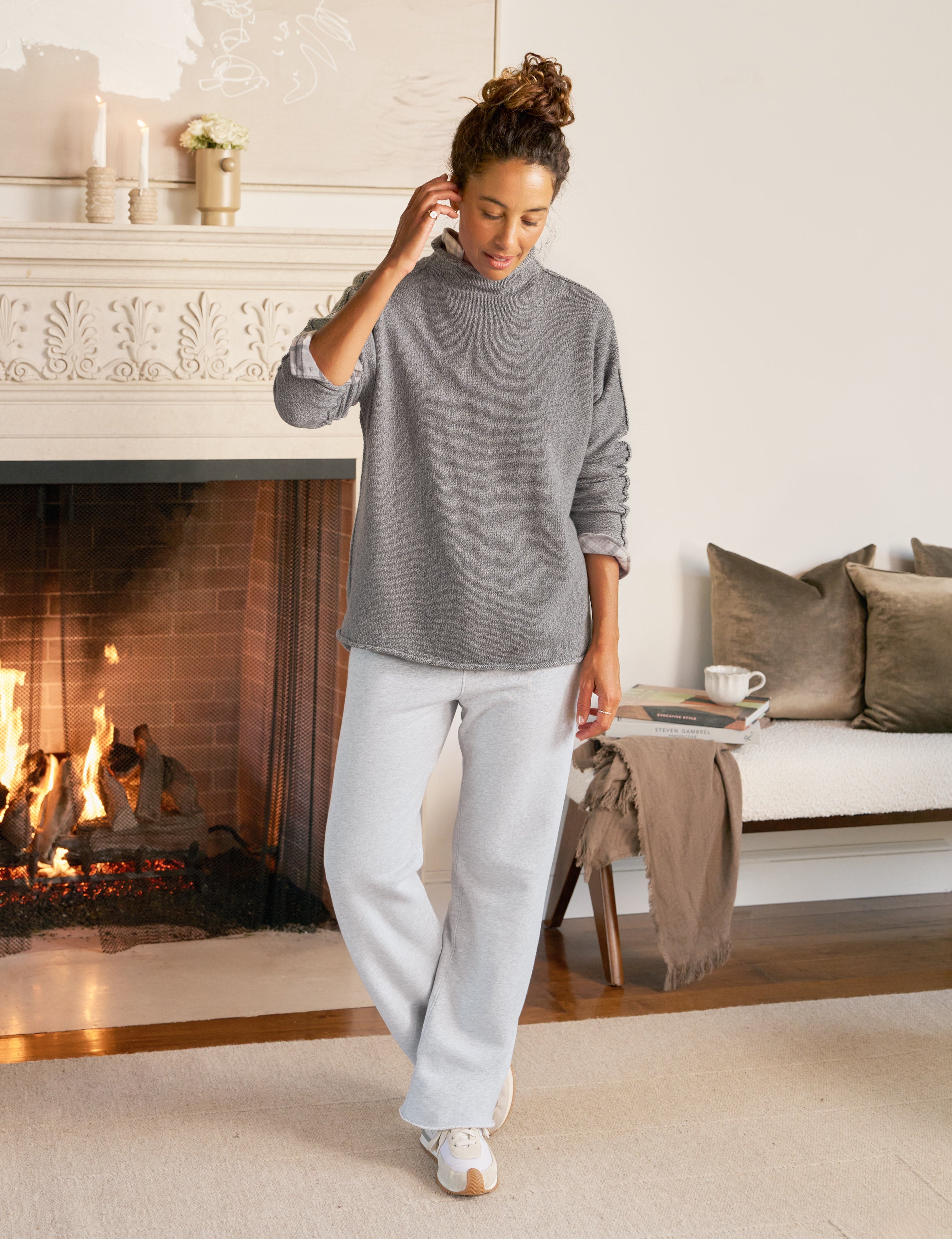 Woman in gray sweater and light gray pants standing in a cozy living room with a fireplace.