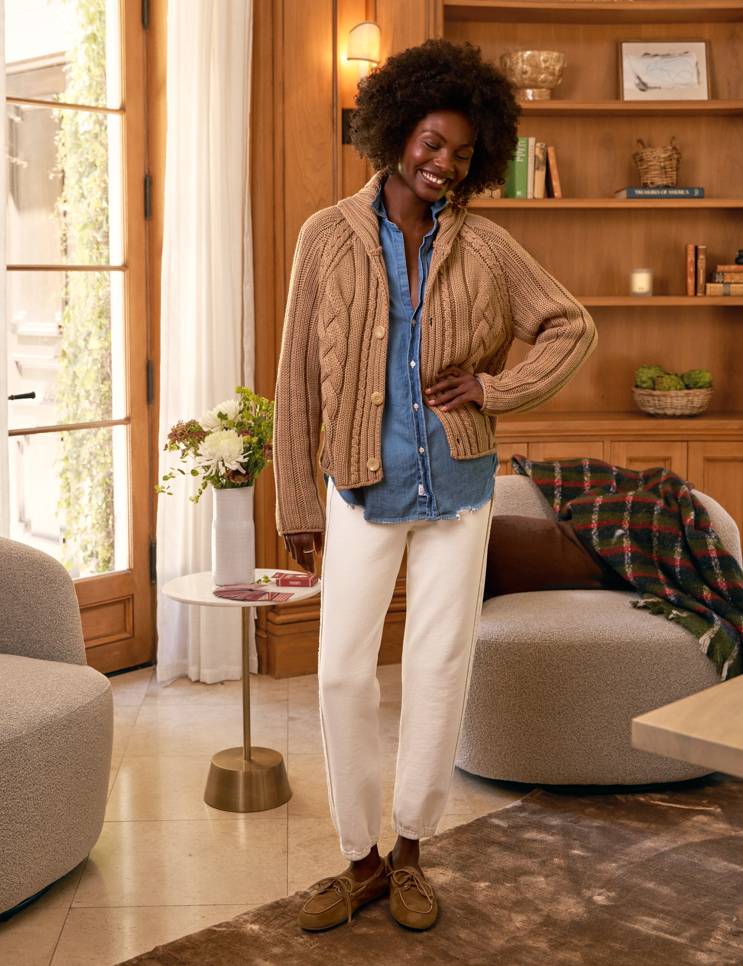 Woman standing in a living room wearing a beige cardigan, blue shirt, and white pants.