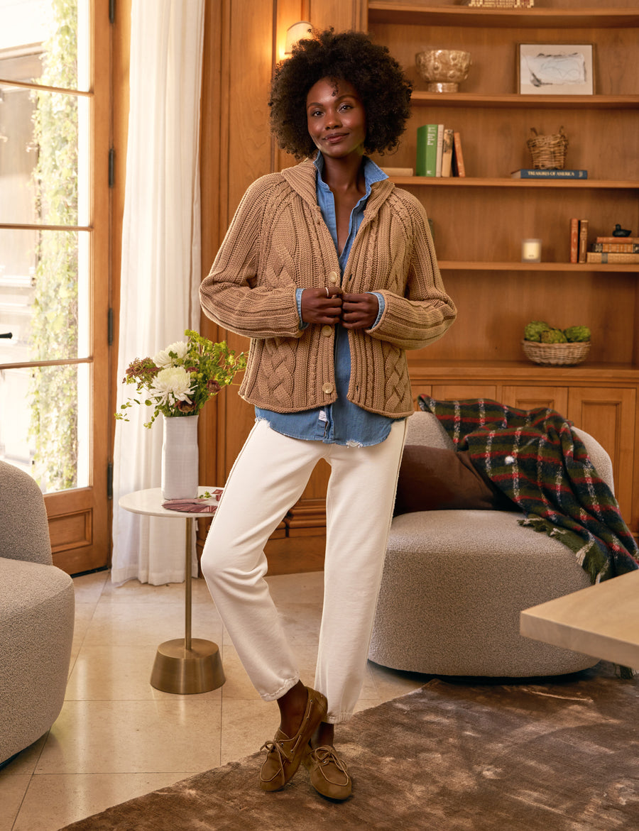 Woman standing in a cozy living room wearing a brown cardigan, blue shirt, and white pants.