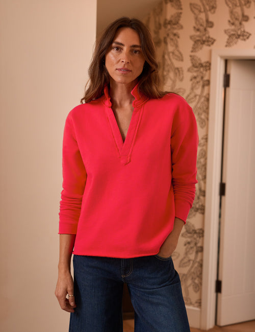 A woman wearing a bright red long-sleeve pullover sweater and dark blue jeans, standing in an indoor setting with patterned wallpaper and a door in the background., front view