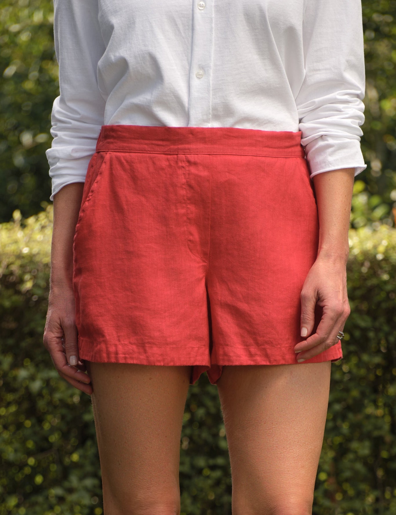 Person wearing red shorts and a white shirt standing on grass with a hedge in the background, front view