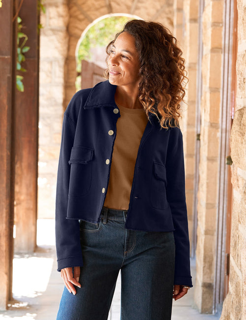 A woman stands in a sunlit stone corridor wearing a navy cropped jacket over a tan top, blue wide-leg jeans, and tan loafers., front view