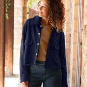 A woman stands in a sunlit stone corridor wearing a navy cropped jacket over a tan top, blue wide-leg jeans, and tan loafers.