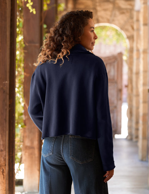 A woman with curly brown hair is shown from behind wearing a navy blue cropped long-sleeve jacket and blue jeans, standing in a stone archway corridor., alternate view