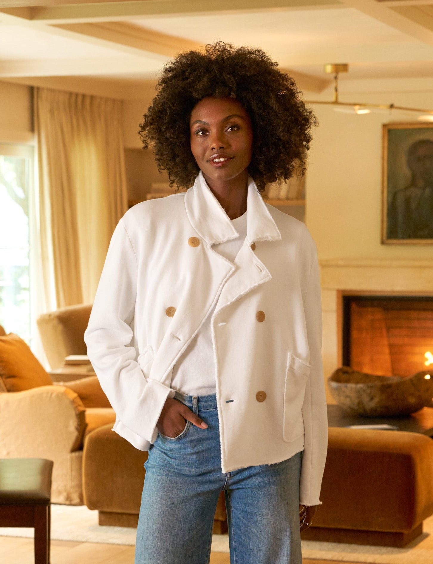 Woman standing in a living room wearing a white coat and blue jeans.