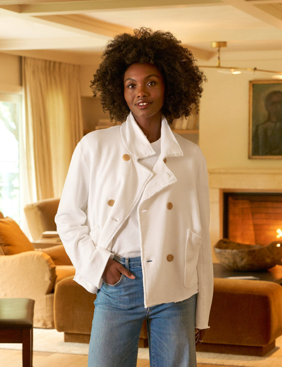 Woman standing in a living room wearing a white coat and blue jeans.