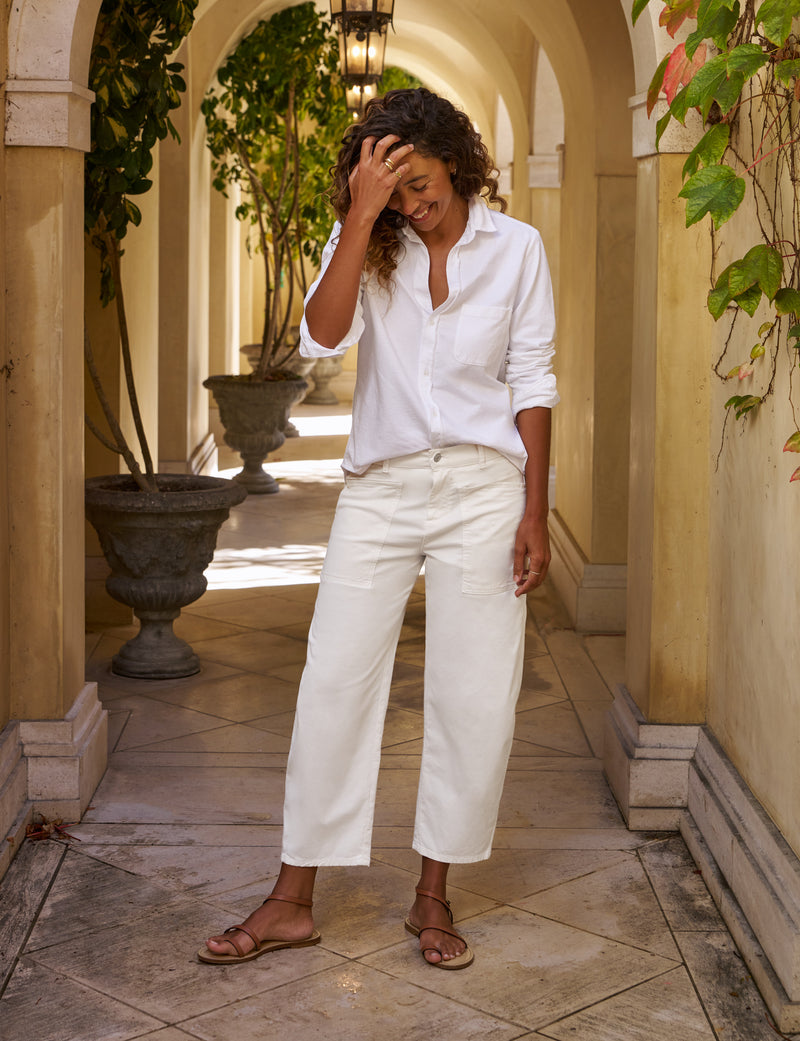 Woman in a white shirt and pants standing in a sunlit hallway with columns and plants.