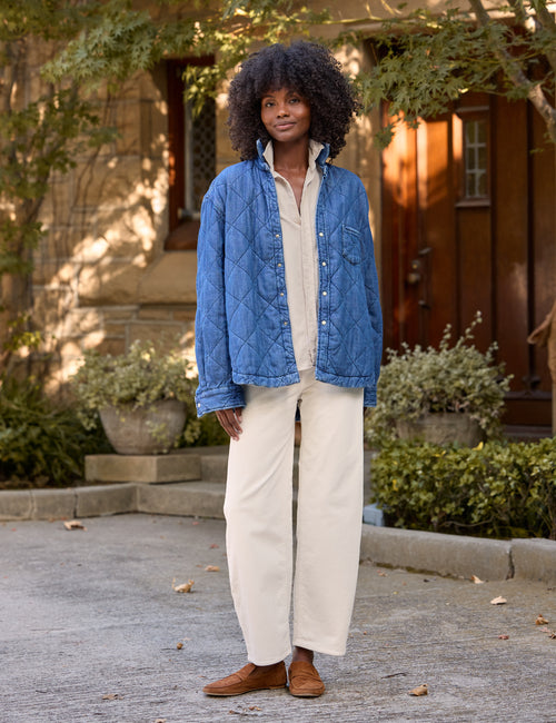 A woman stands outdoors in front of a stone building wearing a blue quilted jacket over a cream button-up shirt, cream barrel-leg pants, and brown shoes., view 6