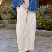 A person standing outdoors in front of a stone building and trees, wearing a blue quilted button-front jacket over a cream shirt and cream wide-leg pants, with brown shoes.