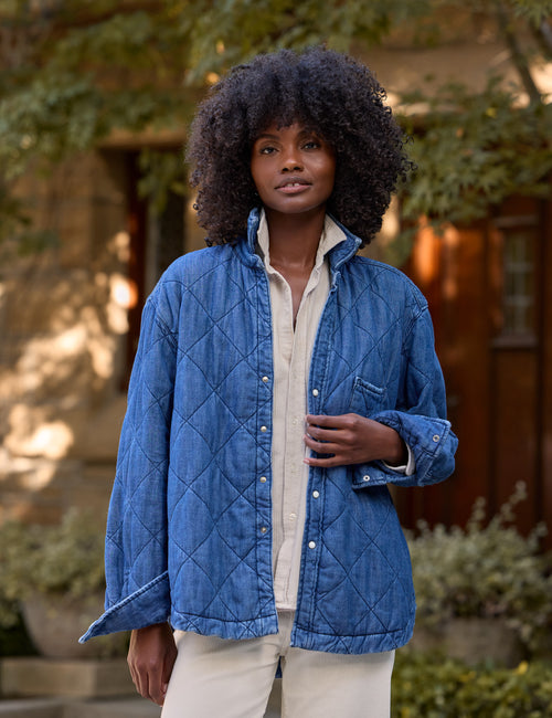 A woman stands outdoors wearing a blue diamond-quilted button-up jacket over a cream shirt and cream pants. The background shows a softly blurred stone building and greenery., front view