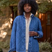 A woman stands outdoors wearing a blue diamond-quilted button-up jacket over a cream shirt and cream pants. The background shows a softly blurred stone building and greenery.