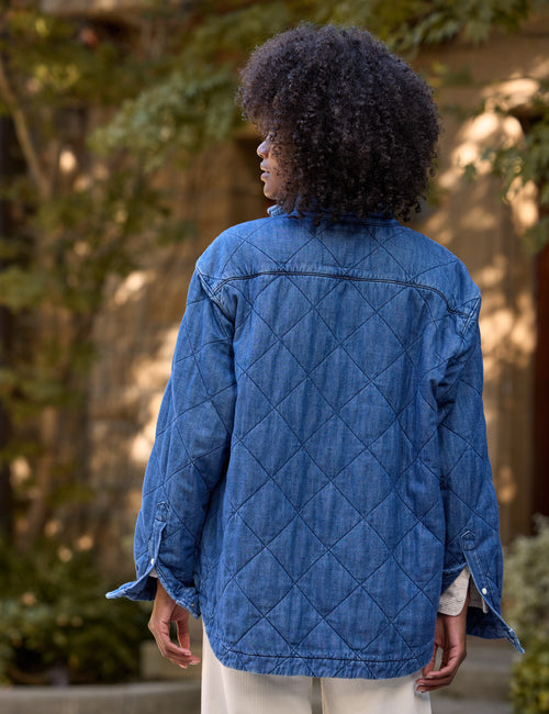 A person viewed from behind wearing a blue quilted jacket with a diamond pattern, set against a blurred outdoor background of green foliage and a stone wall., alternate view