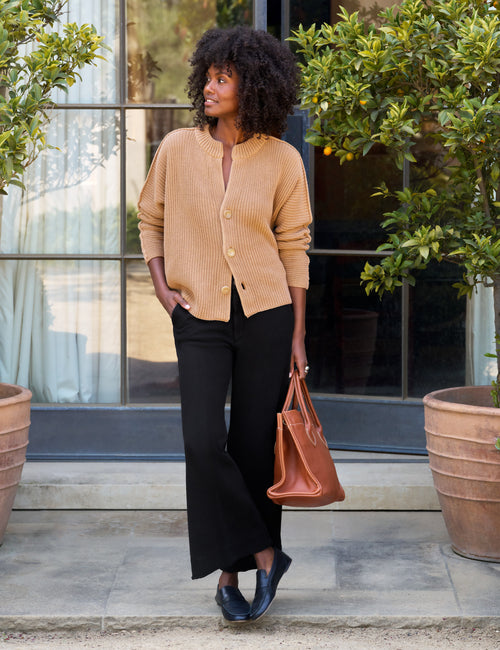 A woman stands outdoors in front of a glass door framed by potted green plants, wearing a tan ribbed cardigan, black wide-leg pants, and black loafers, and carrying a brown leather tote bag., view 5