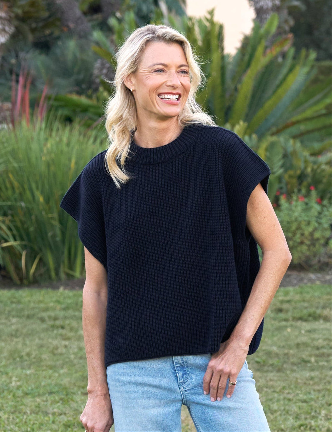 Woman in navy top and light blue jeans holding a hat in a garden setting