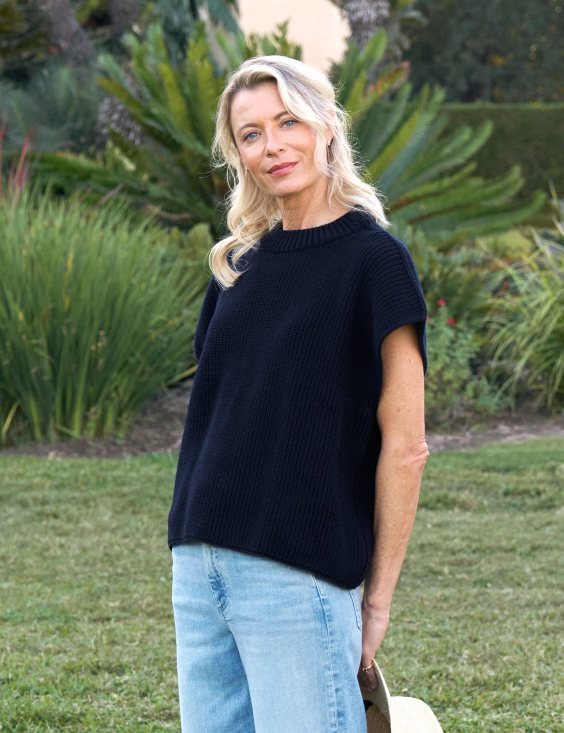 Woman standing on grass wearing a navy top and light blue jeans, holding a beige bag.